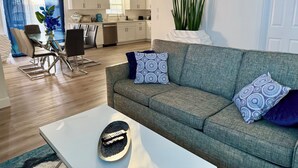 A modern living room with a grey sofa, patterned cushions, a coffee table, and a dining area with chairs and a glass table. - Modern Getaway w/ Roku TVs in Every Room Near Beach + Downtown (St. Petersburg)