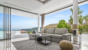 A modern living room with a large sofa, a coffee table, and a view of a beach and mountains. - Jasmine Bophut - Stunning Seaview Villa (Koh Samui)