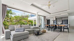 A modern living room with a grey sofa, a glass coffee table, and a dining area with a wooden table and chairs. - Jasmine Bophut - Stunning Seaview Villa (Koh Samui)