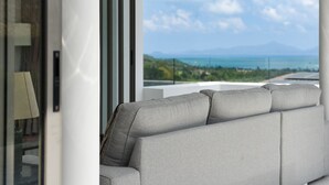 A modern living room with a grey sofa, large windows, and a view of the sea and mountains. - Jasmine Bophut - Stunning Seaview Villa (Koh Samui)