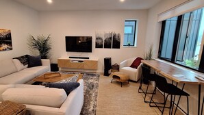 A modern living room with a sofa, coffee table, TV, and a dining area with a window. - The Orange Bridge Lookout | Modern 2BR | Walk to Everything (Nelson)