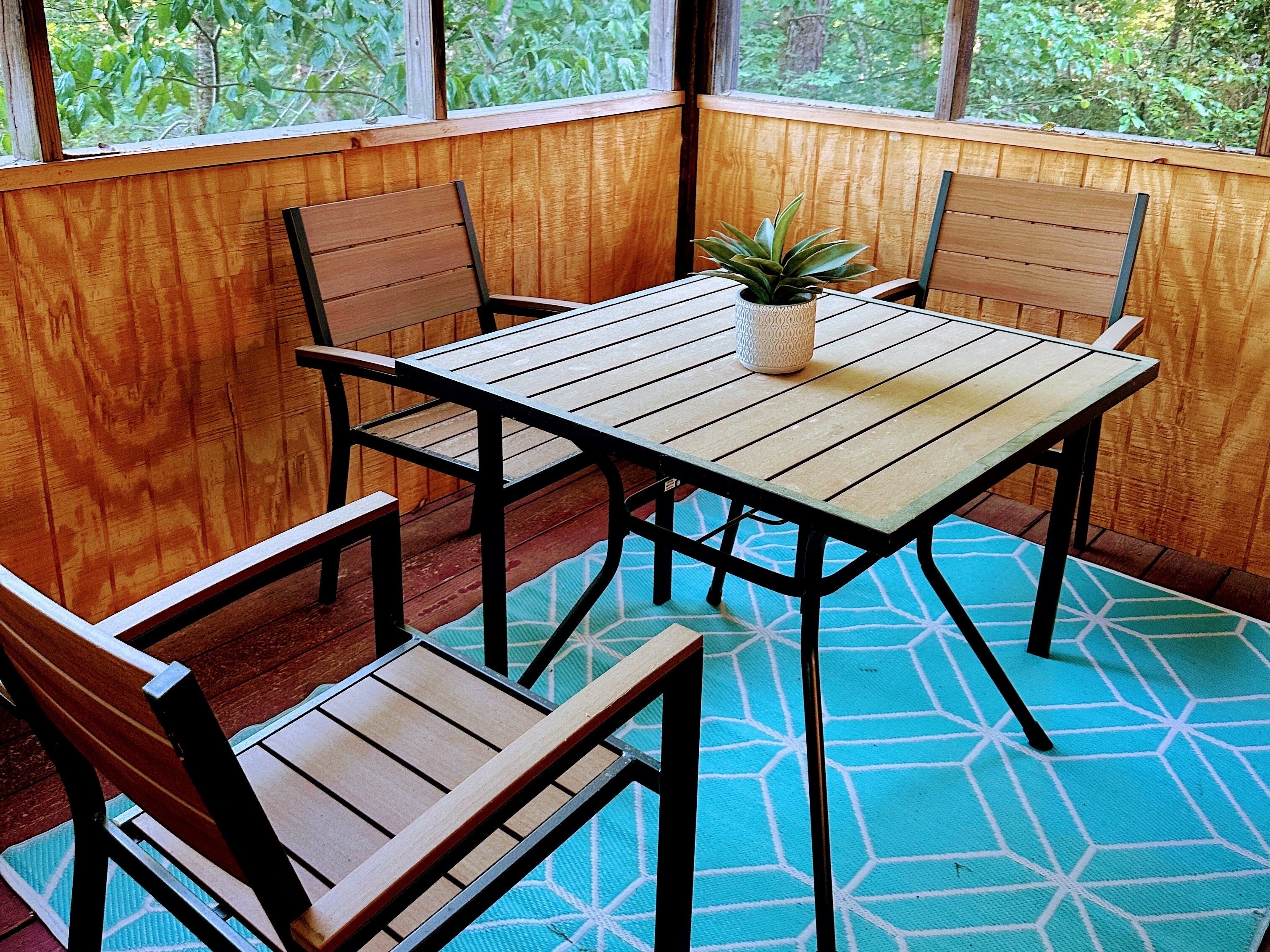 Une table en bois avec des chaises, une plante en pot et un tapis bleu à motifs dans une pièce aux murs en bois et aux grandes fenêtres.