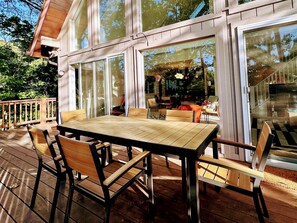 An outdoor deck with a wooden table and chairs, large glass doors leading to an interior space, and a wooden railing. - Lakefront A-Frame: Private Dock, Kayaks & Sauna – Sleeps 12, Chesapeake Bay (Lusby)