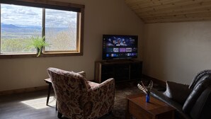 Una sala de estar con televisor, un sillón floral, un sofá de cuero y vistas a montañas a través de un ventanal grande.