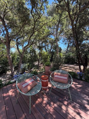A wooden deck with two green metal chairs and a round table, surrounded by trees and potted plants.