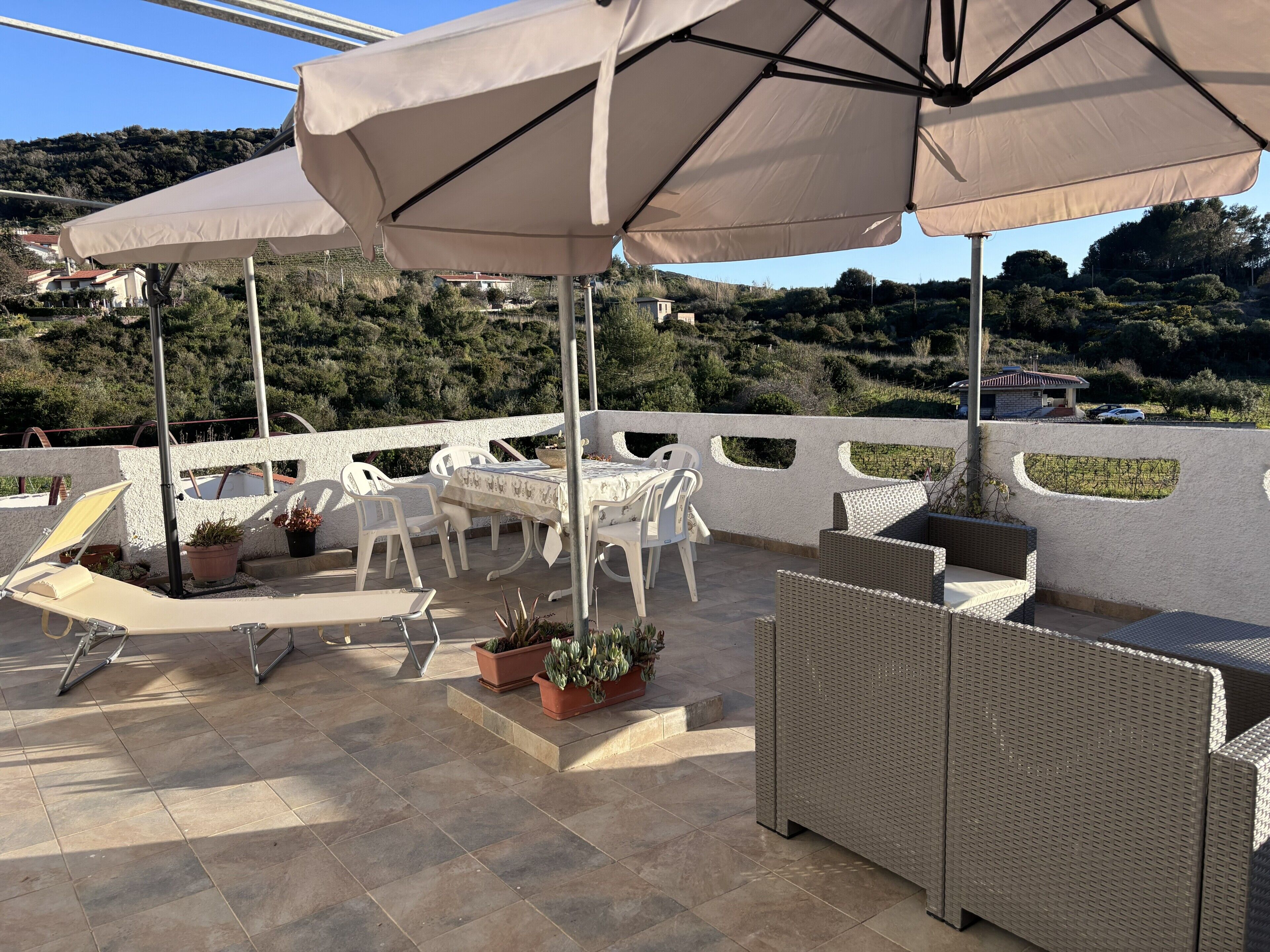 Un patio al aire libre con sillas y mesas de plástico blanco, rodeado de sombrillas beige, plantas en macetas y vistas a un paisaje montañoso.
