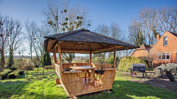 Um gazebo de madeira com uma mesa e bancos, cercado por um gramado e árvores.