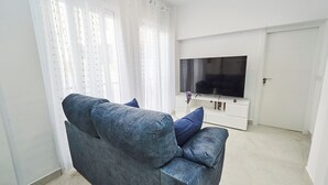 Un salon avec un canapé gris, un téléviseur à écran plat posé sur un meuble blanc, et une porte visible au fond.