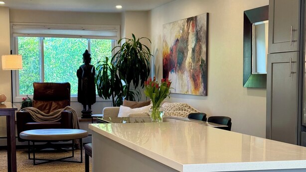 Een moderne keuken met een centraal eiland, een zithoek met een bank en stoelen, een groot raam met uitzicht op groen, en een kleurrijk abstract schilderij aan de muur.