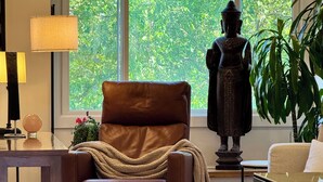 Een moderne woonkamer met een leren fauteuil, een ronde tafel, een beeld en een groot raam met uitzicht op groen.