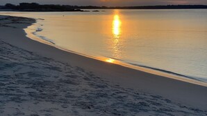 Una playa con huellas que conducen al agua, un mar tranquilo y una puesta de sol al fondo.