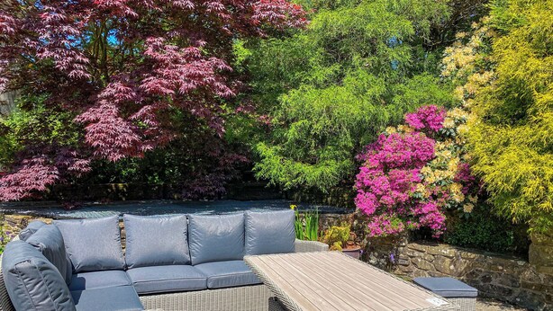 A wicker outdoor seating area with cushions and a table, surrounded by lush greenery and flowering plants. - Dyffryn Conin is more than just a place to stay. (Clynderwen)