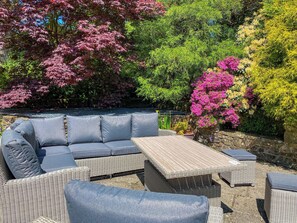 A wicker outdoor seating area with cushions and a table, surrounded by lush greenery and flowering plants. - Dyffryn Conin is more than just a place to stay. (Clynderwen)