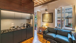A modern kitchen with dark cabinets and a wooden floor, a grey sofa, and a window with a view of buildings. - Delightful Penthouse in the heart of Monti within walking distance metro Cavour and Colosseum. (Roma)
