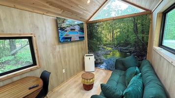Un acogedor interior de cabaña con techo de madera, un sofá verde, una mesita y un televisor que muestra una escena tropical.