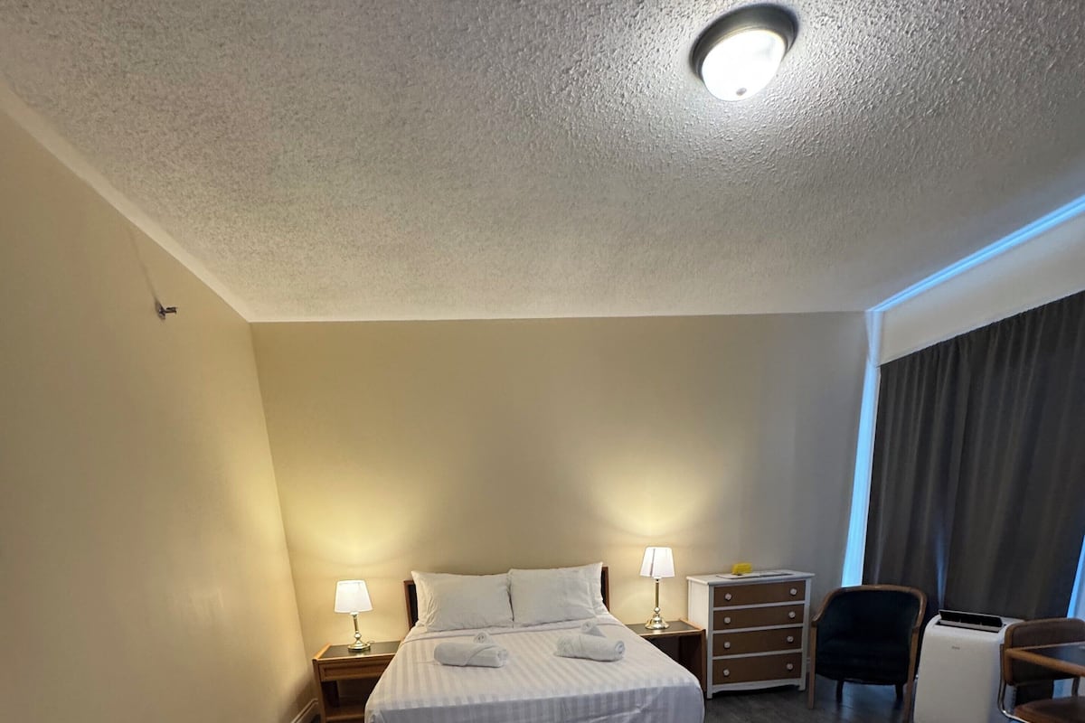 A hotel room with a bed, two bedside tables, a desk, a chair, a lamp, a small table with a yellow object, and a large rug.