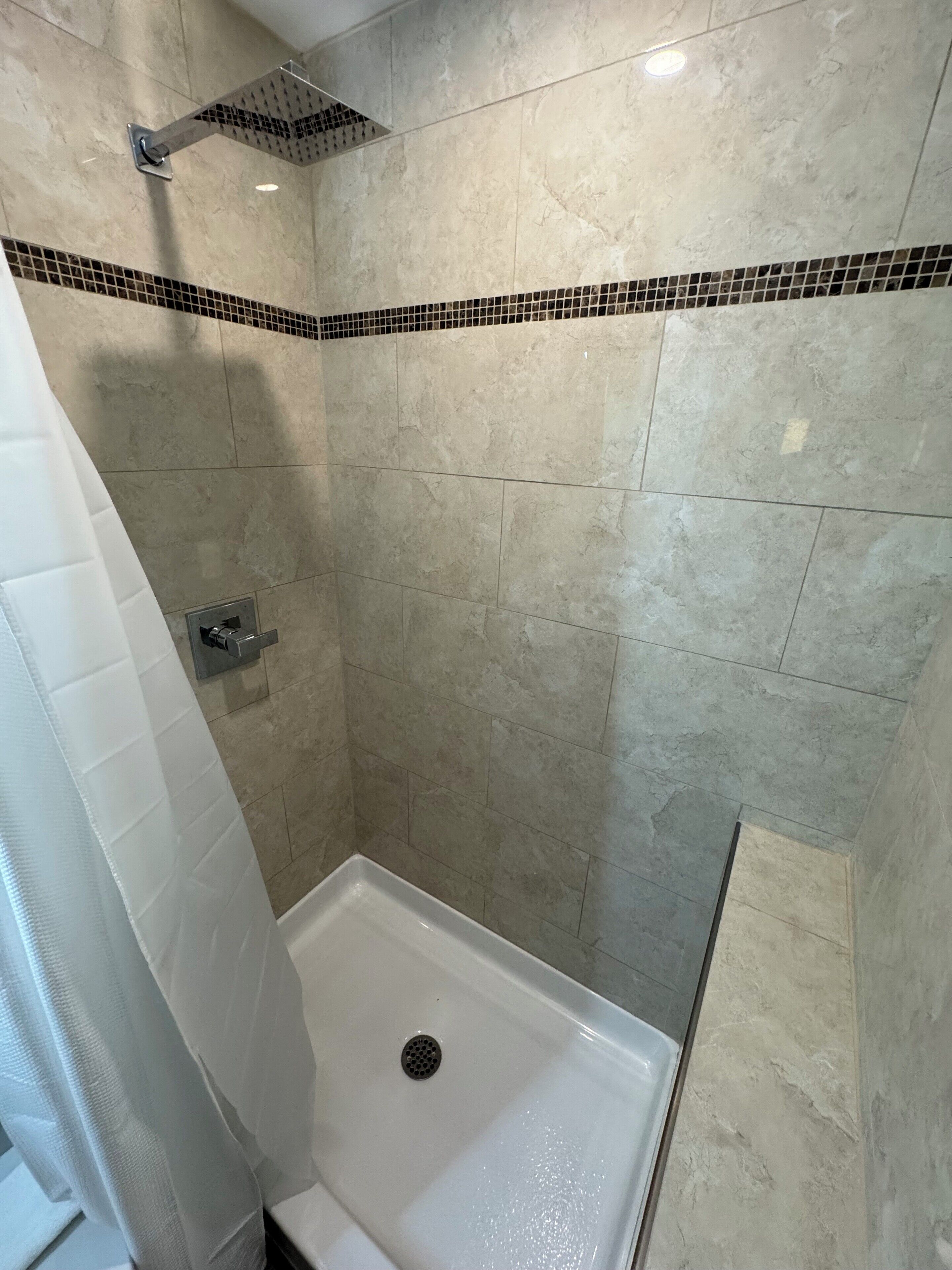 A shower with a square showerhead, a tiled wall, and a white shower curtain.