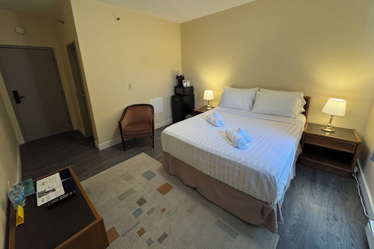 A hotel room with a bed, two bedside tables with lamps, a chair, a desk with a remote control, and a small rug.