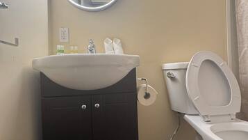A bathroom with a white sink, a mirror with a light, a toilet, and a trash bin.