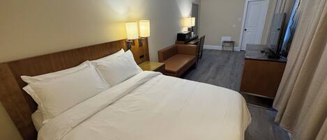 Una habitación de hotel con una cama grande, un escritorio con silla, un televisor y una mesita de noche con lámpara.