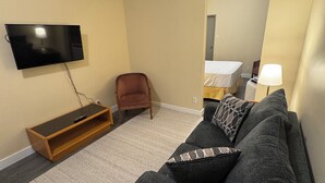 A living room with a flat-screen TV mounted on the wall, a wooden TV stand, a grey sofa with patterned cushions, a brown armchair, and a bed with a white mattress and patterned blanket in the background. - Kenricia Hotel (Kenora)