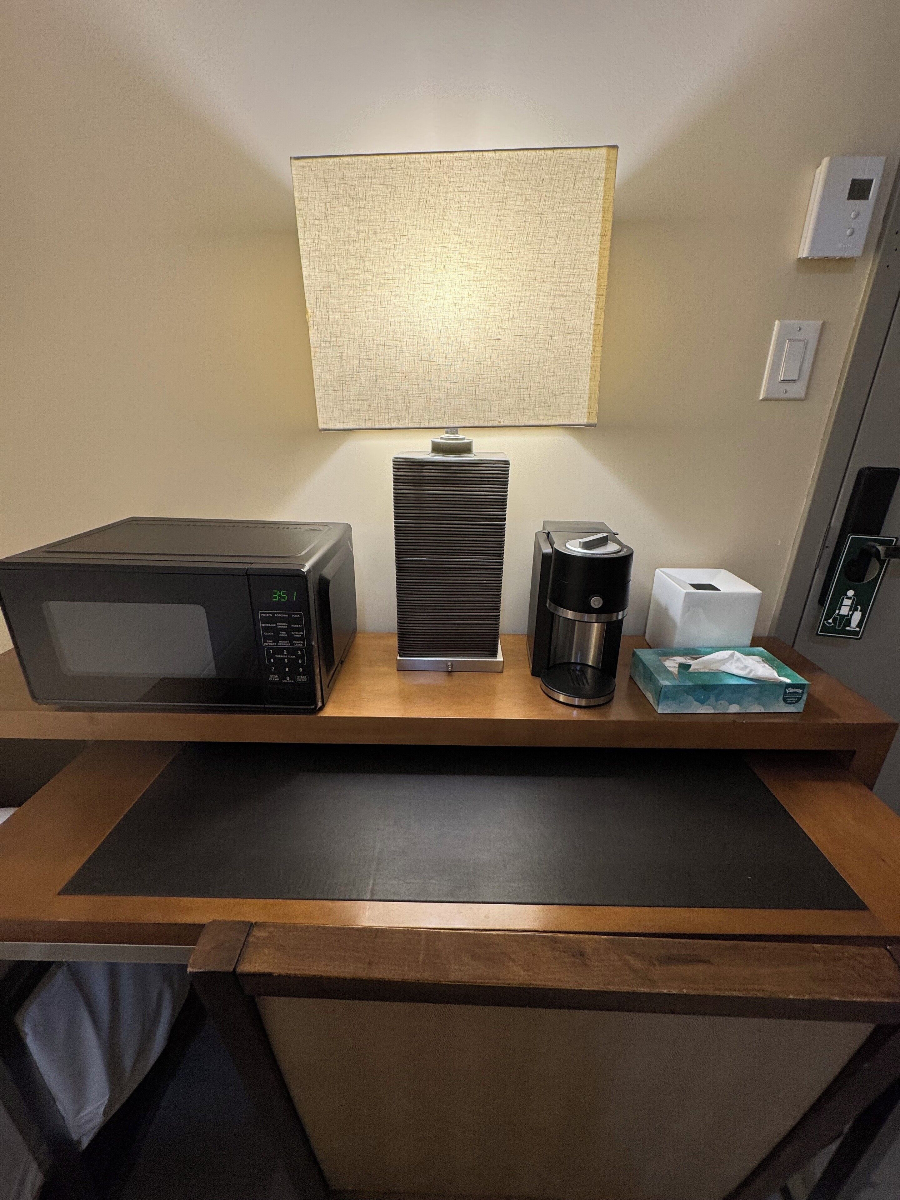 A hotel room desk with a microwave, coffee maker, tissue box, and a lamp.
