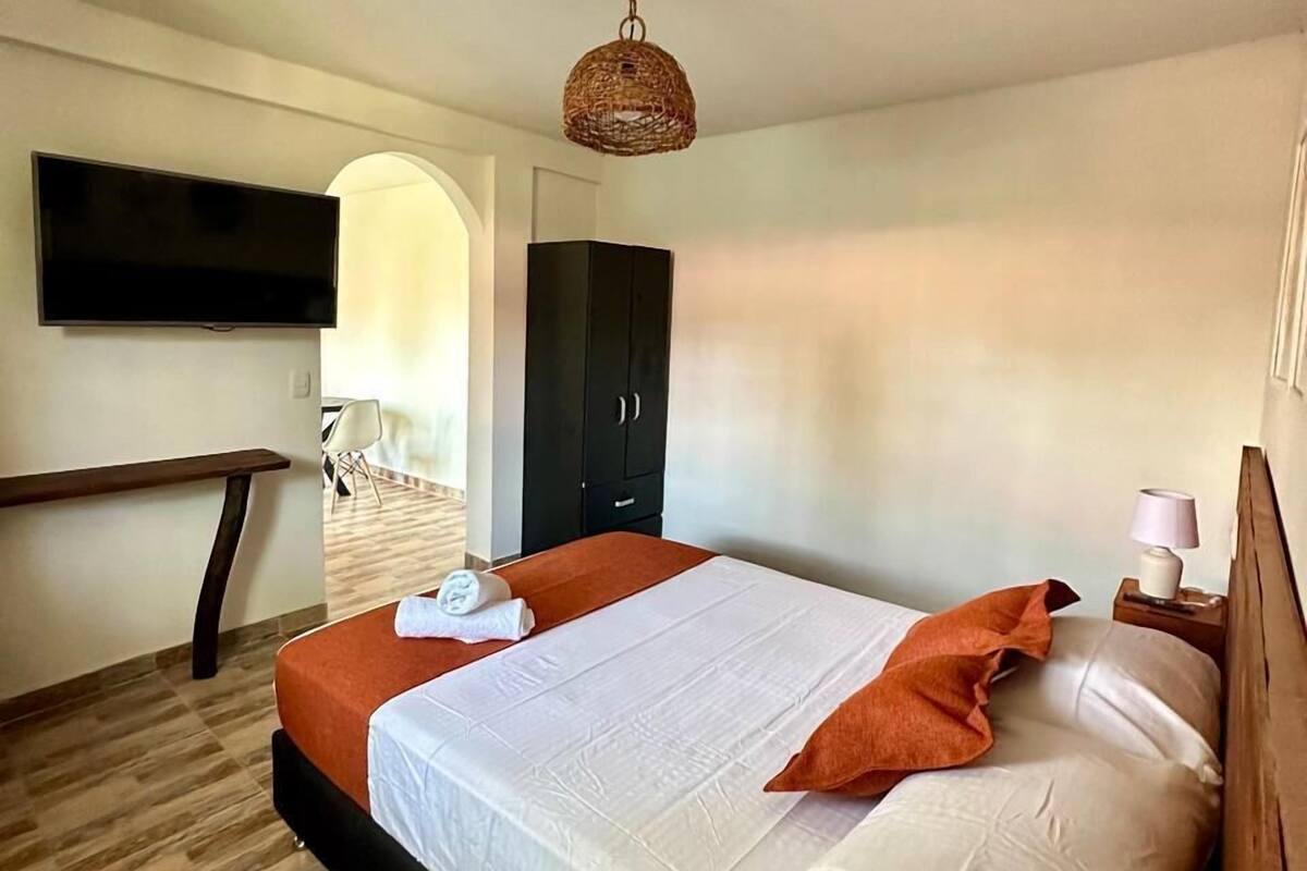 A modern hotel room with a large bed, a flat-screen TV mounted on the wall, a wooden bench, and a wicker ceiling light.