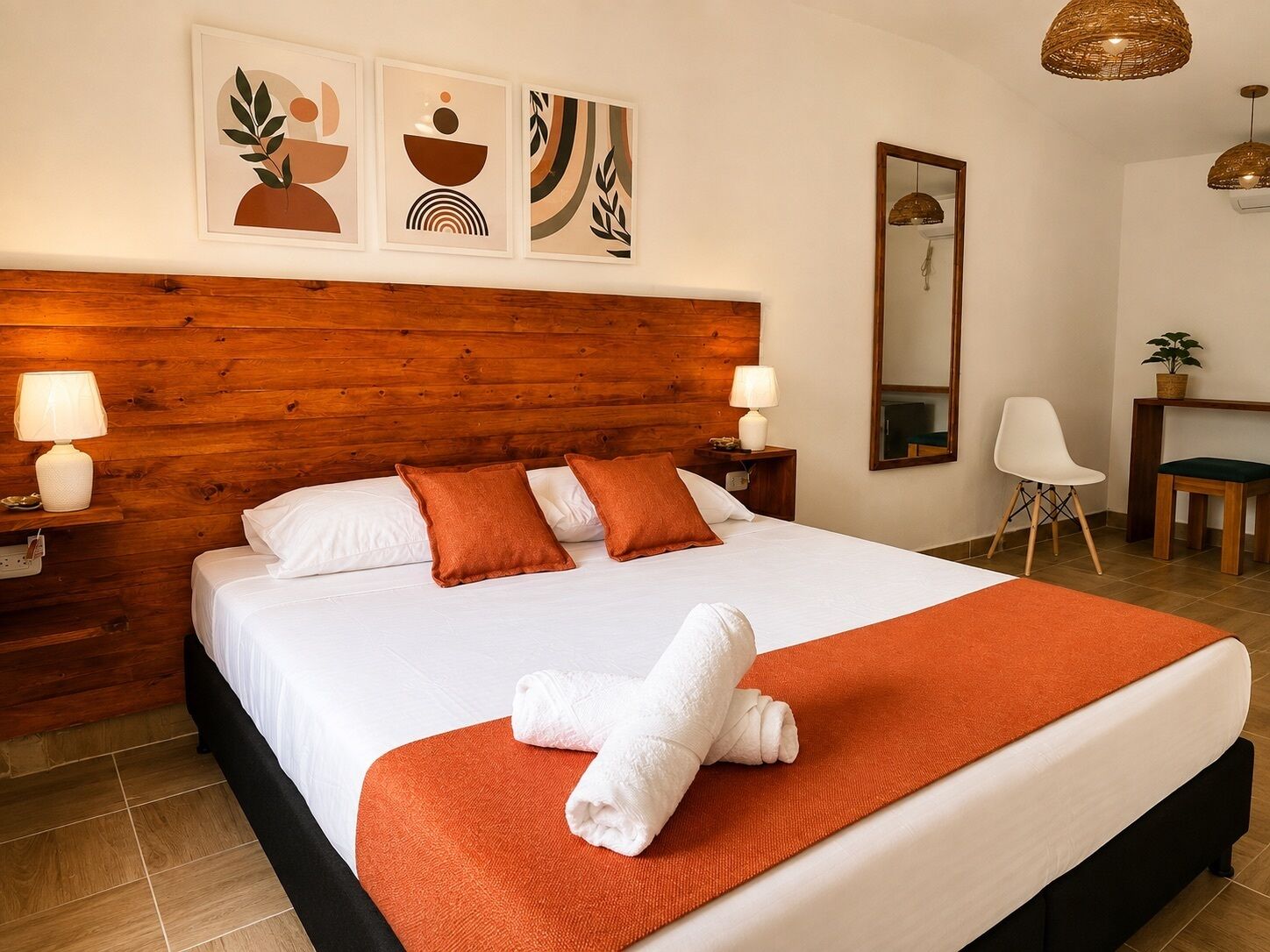 Habitación de hotel con cabecera de madera, cama con ropa blanca y detalles naranja, una toalla blanca sobre la cama, mesita de noche con lámpara, una silla, escritorio con planta y obra de arte en la pared.