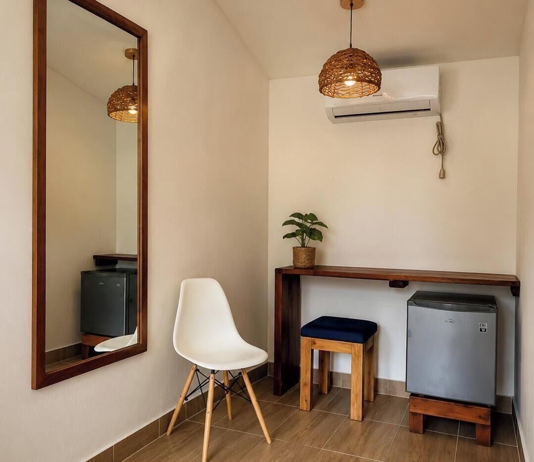 Una habitación compacta con un espejo grande, una silla blanca, un taburete pequeño, una mesa de madera, una planta en maceta y un refrigerador pequeño.