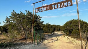 Un segnale con la scritta 'Sueno Texano' in un ambiente rurale, con una strada sterrata e vegetazione rada.