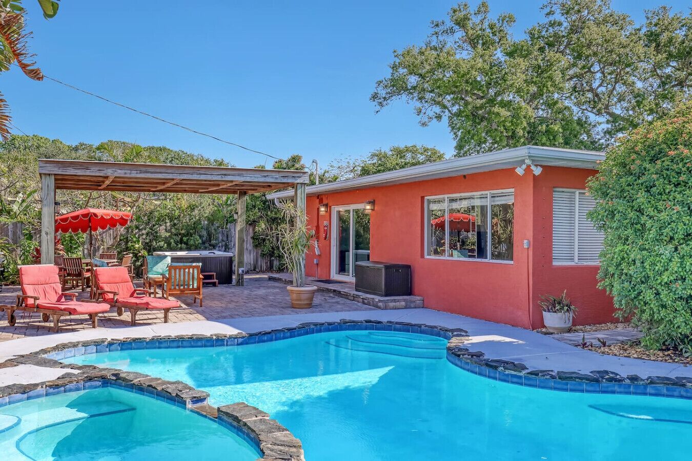 A backyard with a swimming pool, a covered patio with chairs, and a red building.