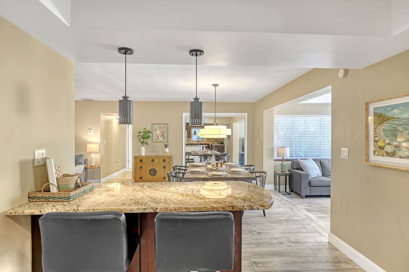 A spacious kitchen with a marble island, pendant lighting, and a dining area with a gray sofa and a painting on the wall.