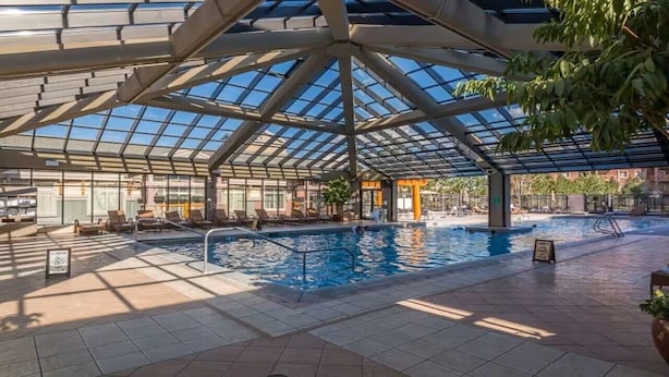 An indoor swimming pool with a glass ceiling, surrounded by lounge chairs and potted plants.