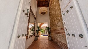 A corridor with white walls, a tiled floor, and a wooden ceiling. There are potted plants and a hanging light fixture. - Casa Bovedas - 5 Bedroom Paradise (Cartagena de Indias)