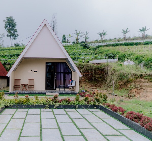 A small, single-story building with a triangular roof, a front porch with two chairs and a table, and a garden area with various plants. - Vianda Cabin By Travelink (Garung)