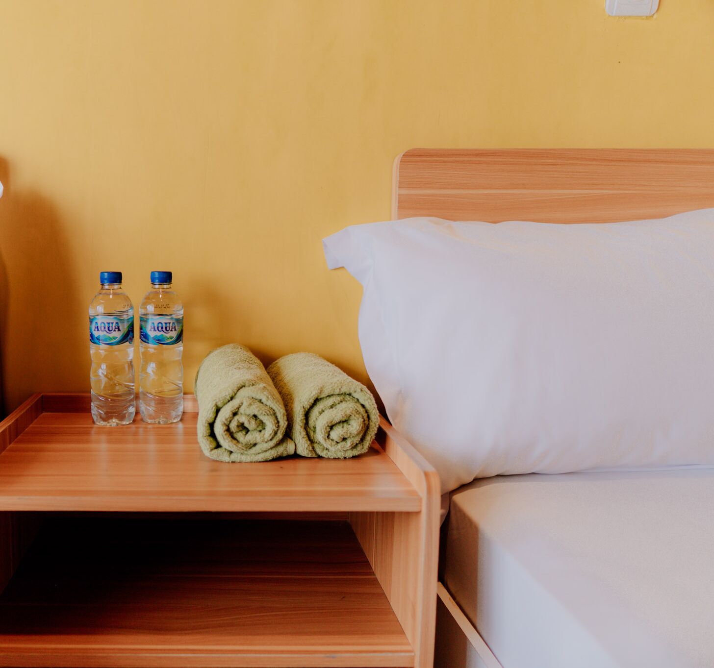 Ein Nachttisch aus Holz mit zwei Wasserflaschen und zwei zusammengerollten Handtüchern neben einem Bett mit weißen Laken.