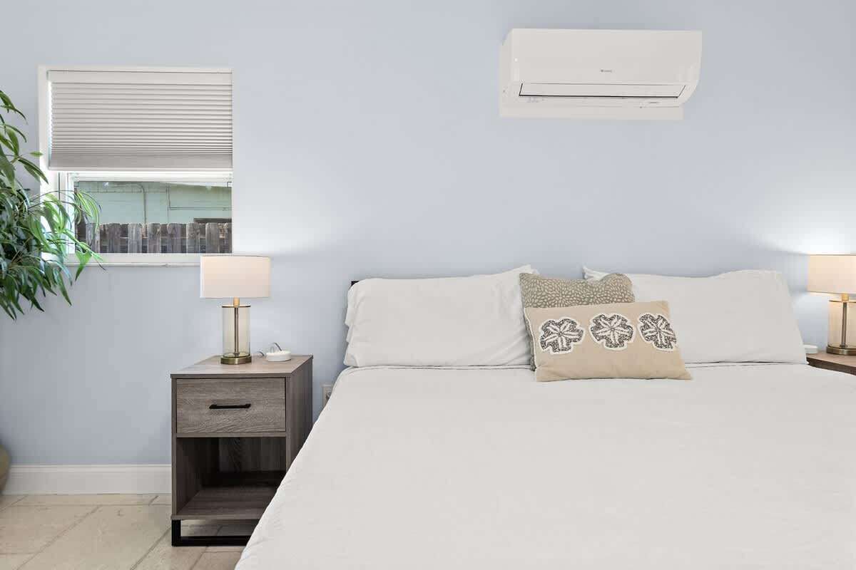 Un dormitorio con una cama, dos mesitas de noche con lámparas, una ventana con persianas y un equipo de aire acondicionado en la pared.