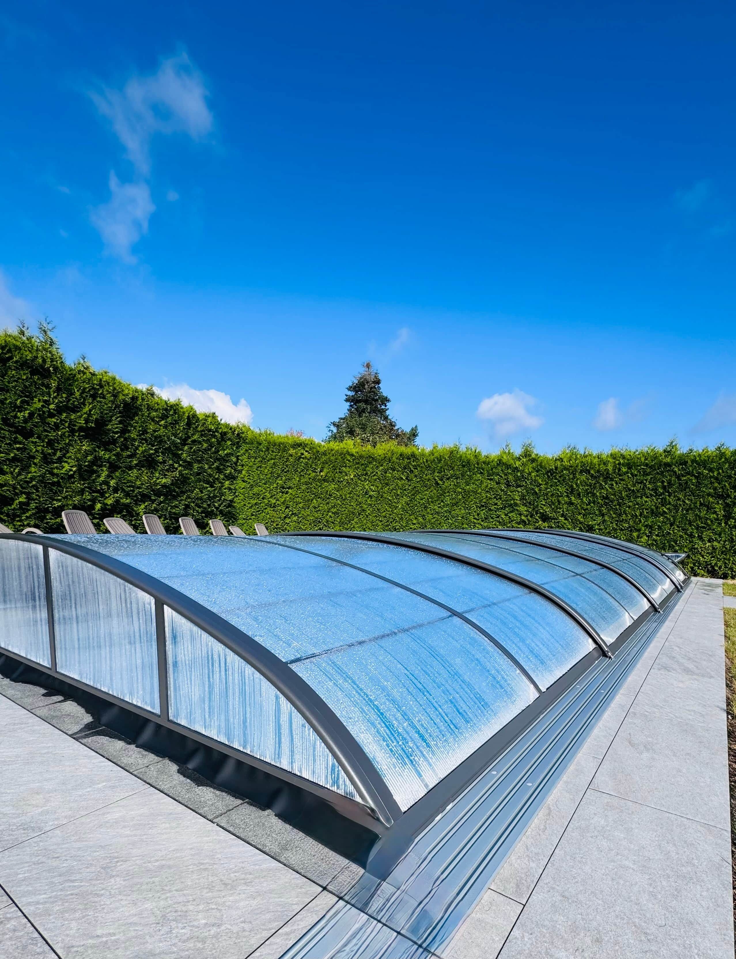 Ein modernes Bauwerk mit einer gewölbten Glasdachkonstruktion, umgeben von einer ordentlich geschnittenen Hecke und blauem Himmel.