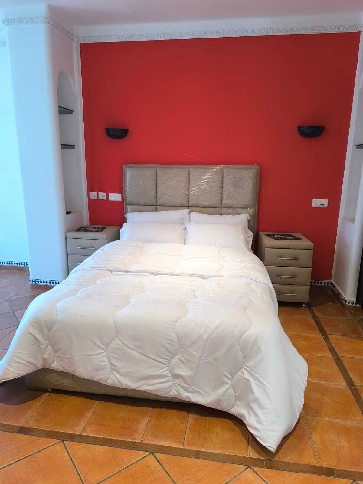 Un lit recouvert d’une couette blanche, dans une pièce aux murs rouges et au sol carrelé.