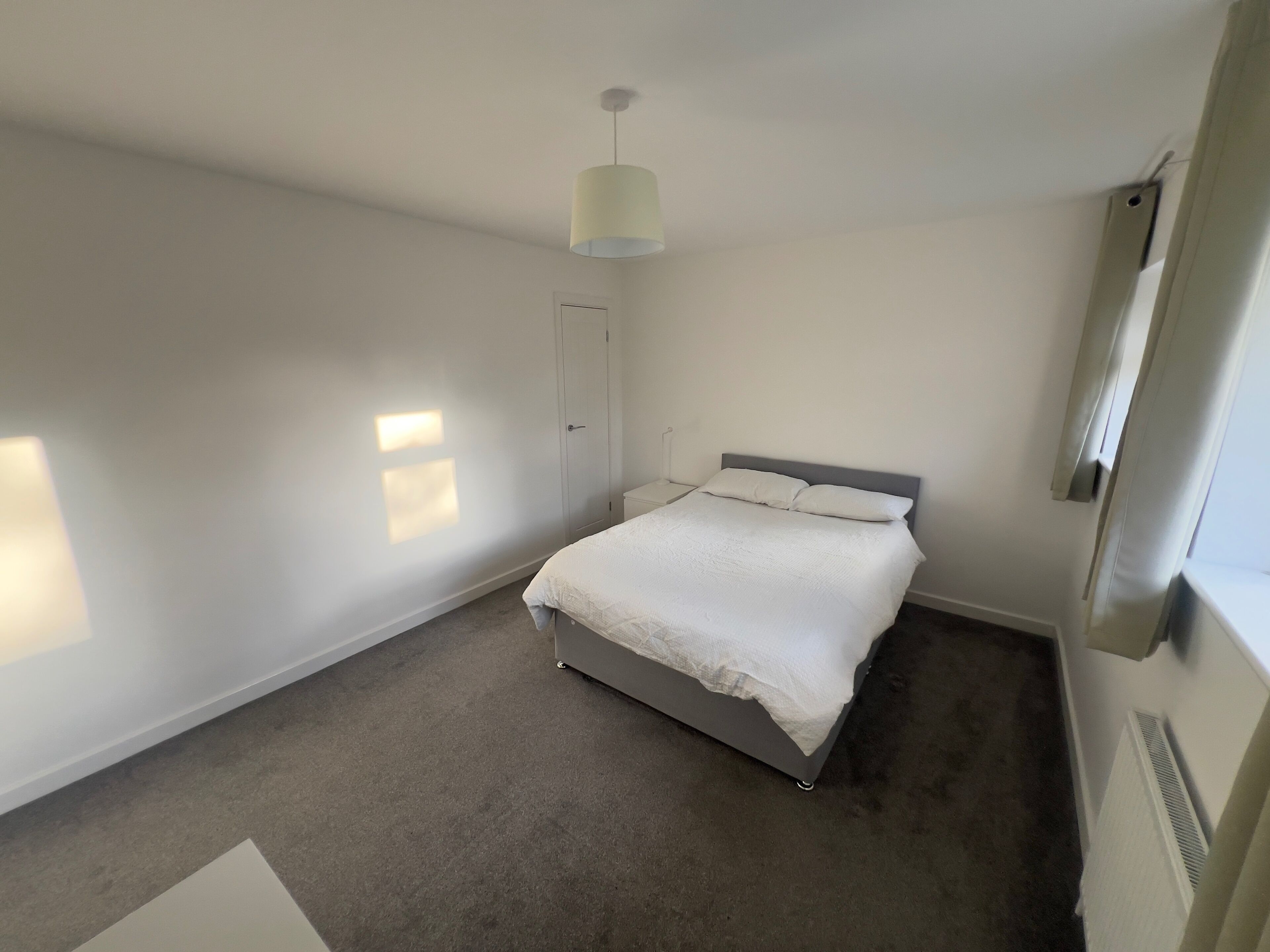 Um quarto com uma cama, uma mesinha de cabeceira, um candeeiro pendente e um radiador.