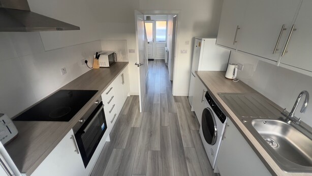 Une cuisine moderne équipée d’un four encastré, d’un micro-ondes et d’un lave-linge. On y trouve un évier et un plan de travail avec un grille-pain. Le sol est en bois et les placards sont blancs.