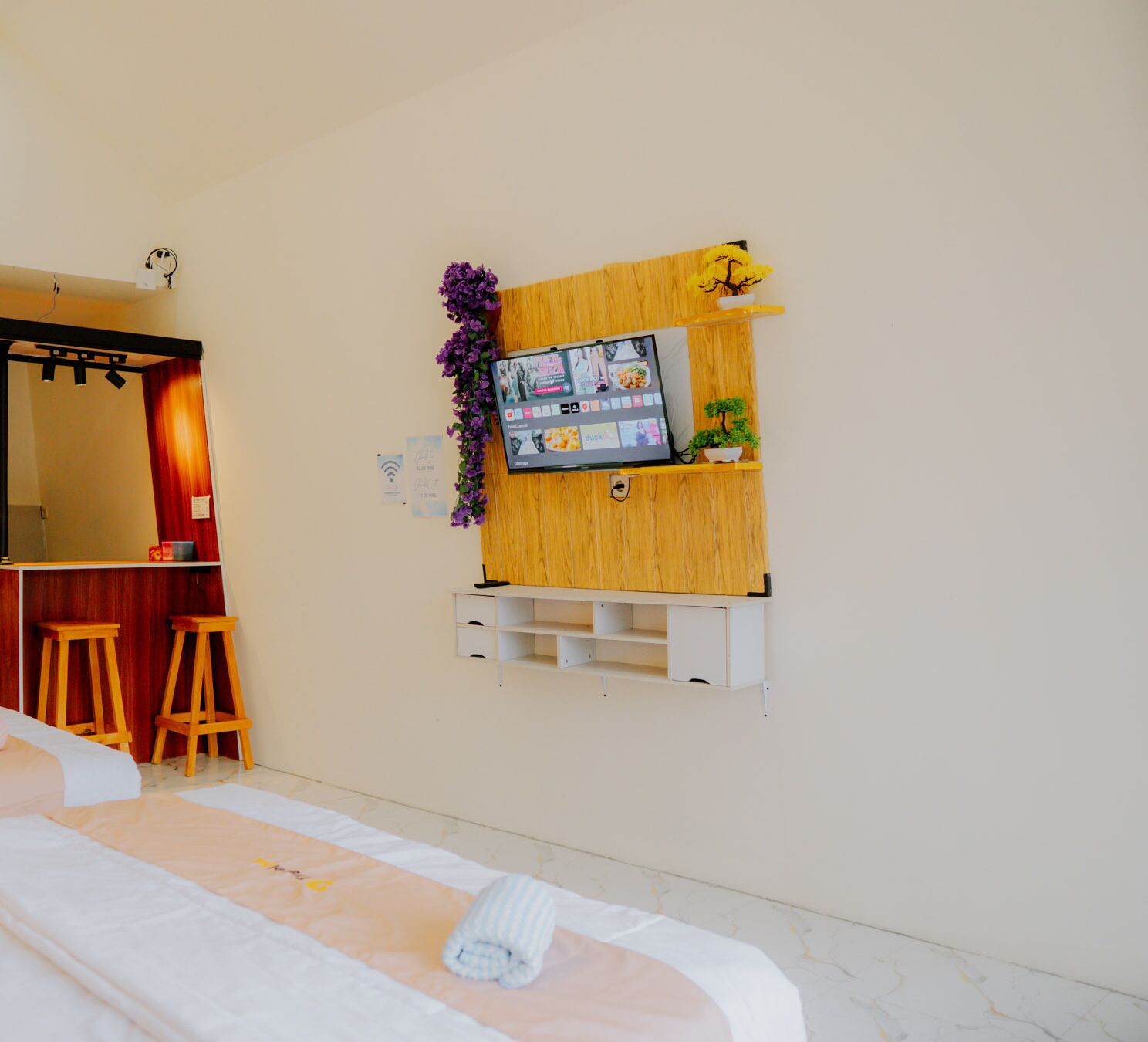 A hotel room with a bed, a wooden shelf with a TV, a small plant, and a purple flower decoration.