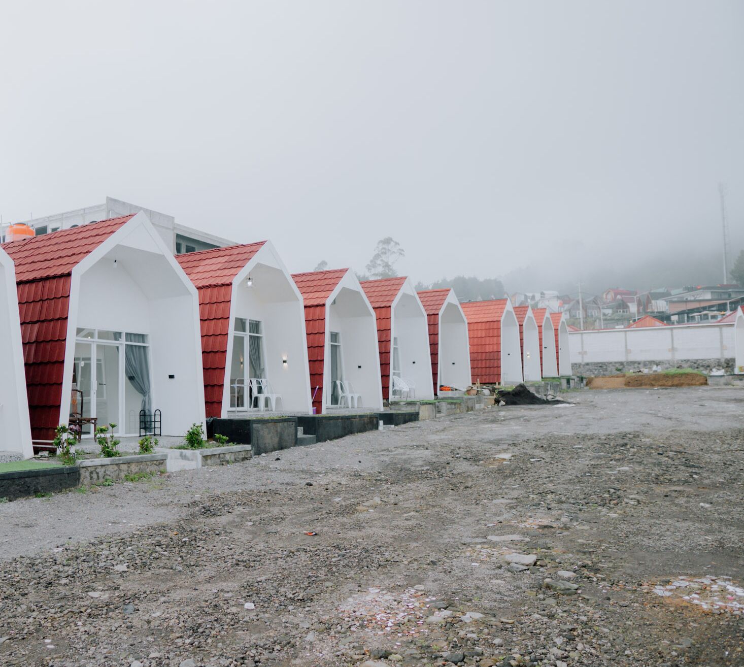 A row of white buildings with red roofs, likely a row of cabins or cottages, on a gravel ground with a foggy background.
