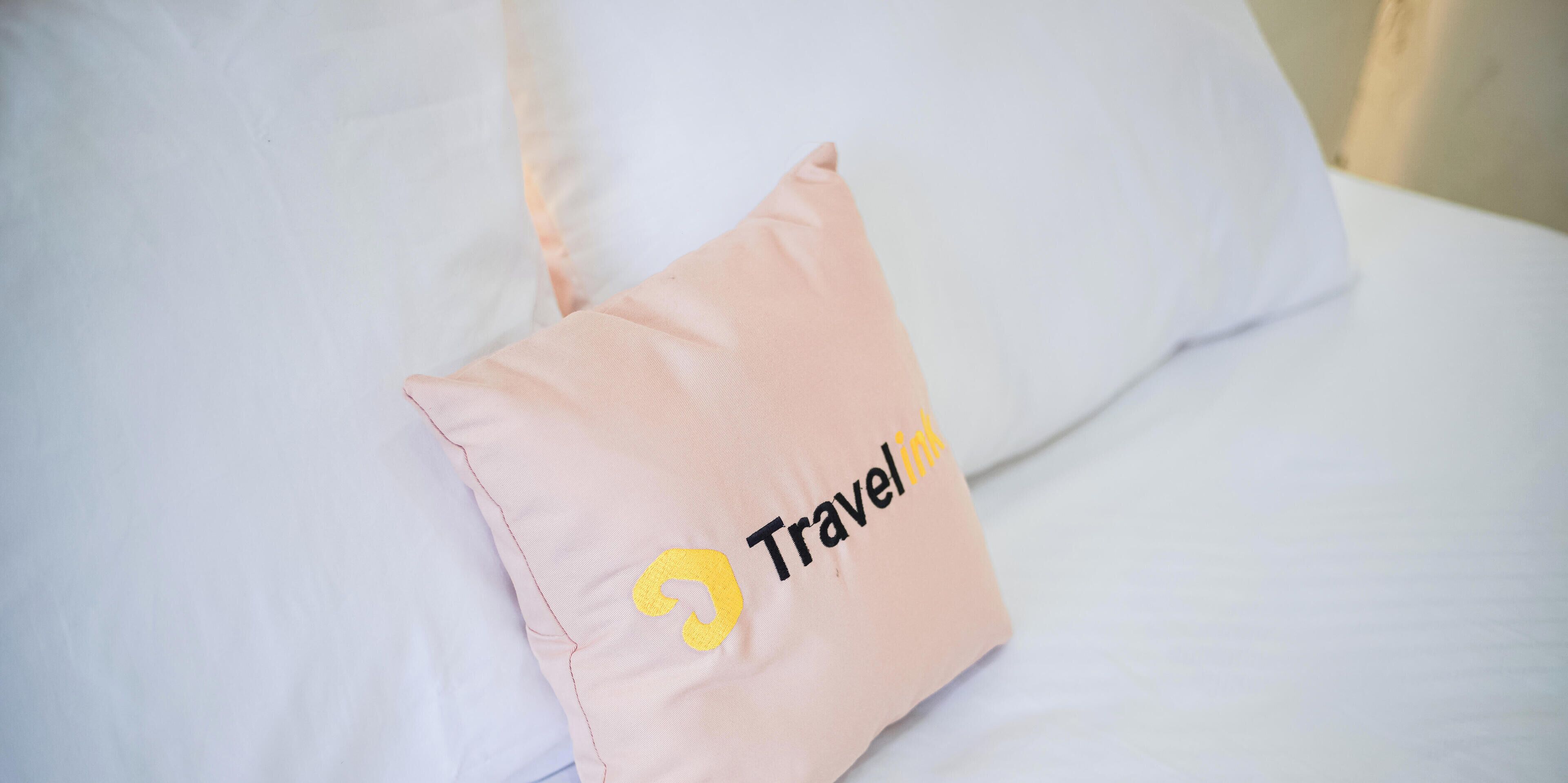 A bed with white bedding and a pink pillow with a logo and text.
