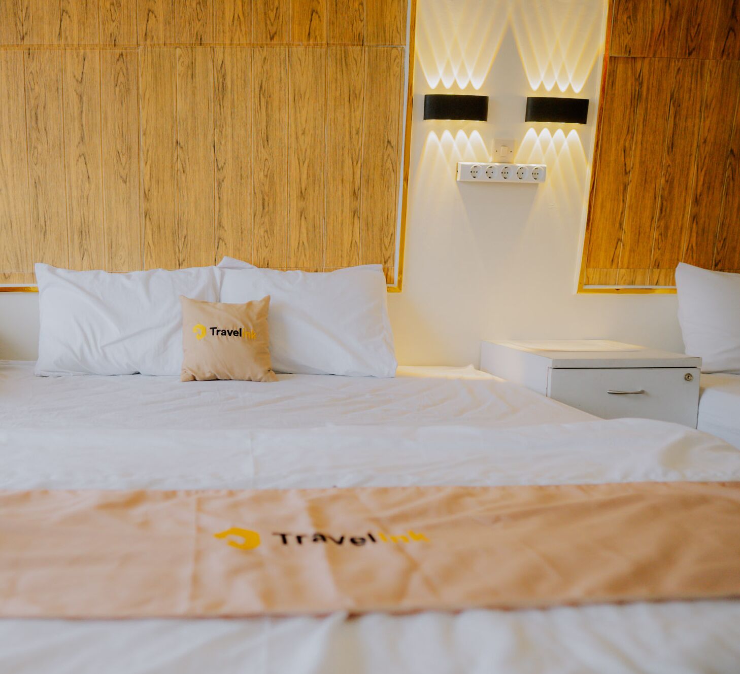A hotel room with a wooden headboard, a bed with white bedding, a nightstand, and a wall-mounted light fixture.