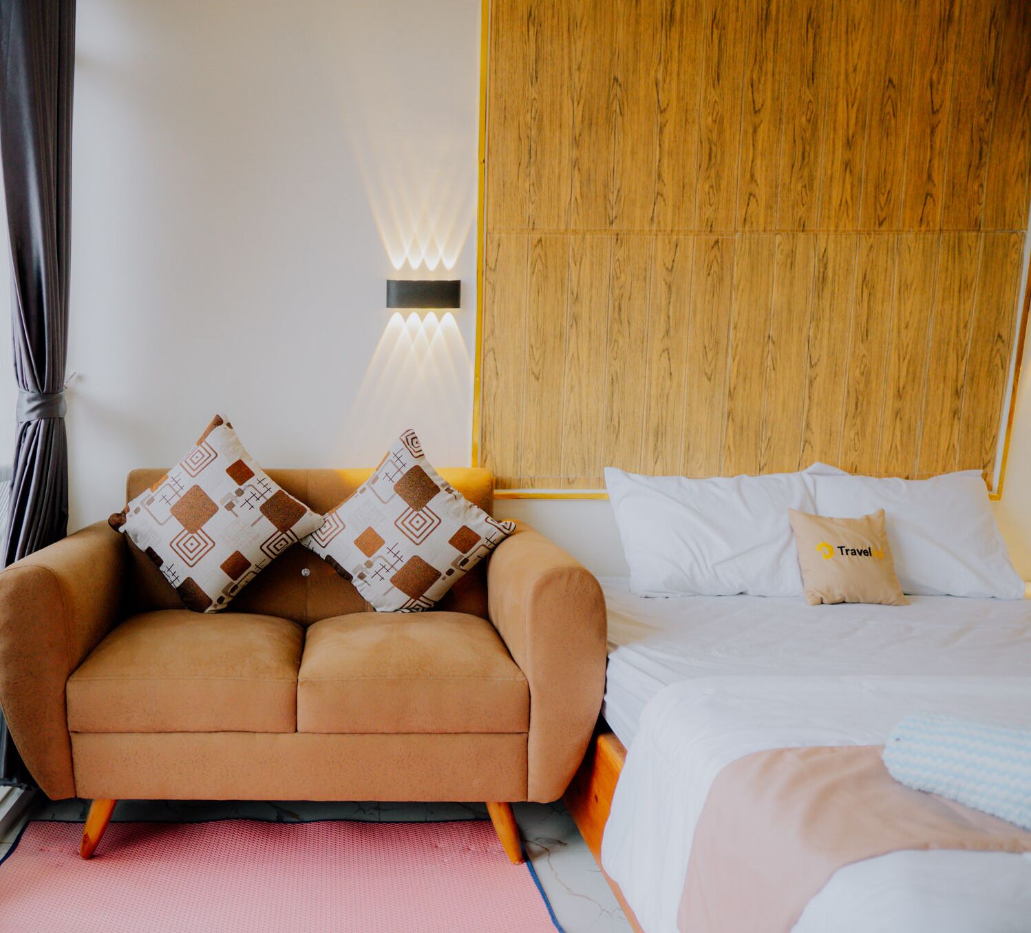 A modern hotel room with a wooden headboard, a brown sofa with patterned cushions, a white bed with a travel pillow, and a red textured rug.