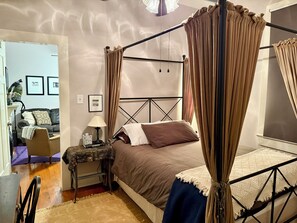 A four-poster bed with a canopy, a bedside table with a lamp, and a chair in a room with a wooden floor.
