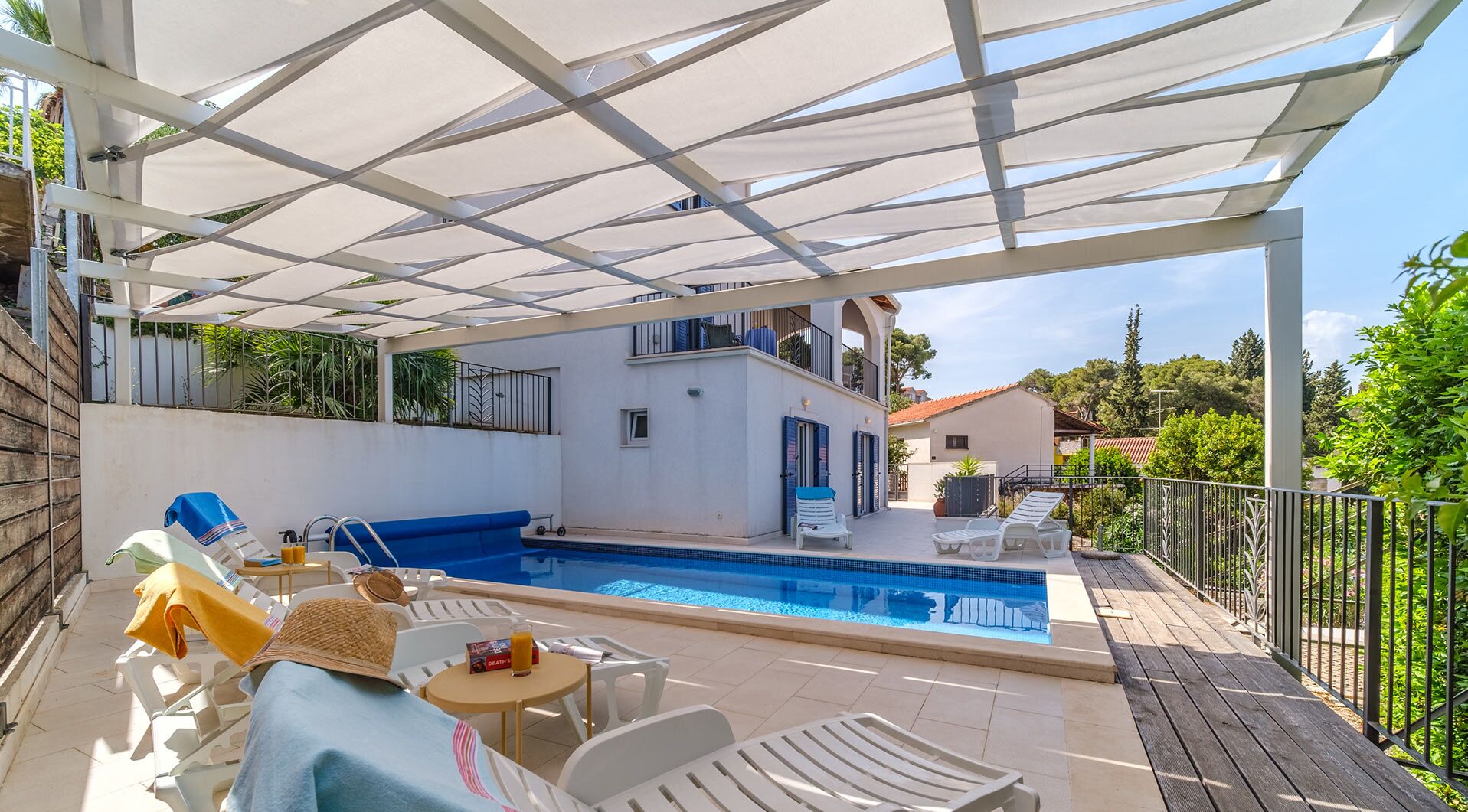 A covered outdoor swimming pool area with lounge chairs and a table, surrounded by a wooden deck and a metal fence.