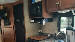 A compact kitchen with a microwave, stove, and sink. There is a trash can and a small window.