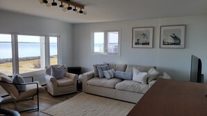 A living room with a sofa, armchair, and a view of the sea through large windows. - Oceanfront cottage with private beach access near Shediac, New Brunswick (Grande-Digue)
